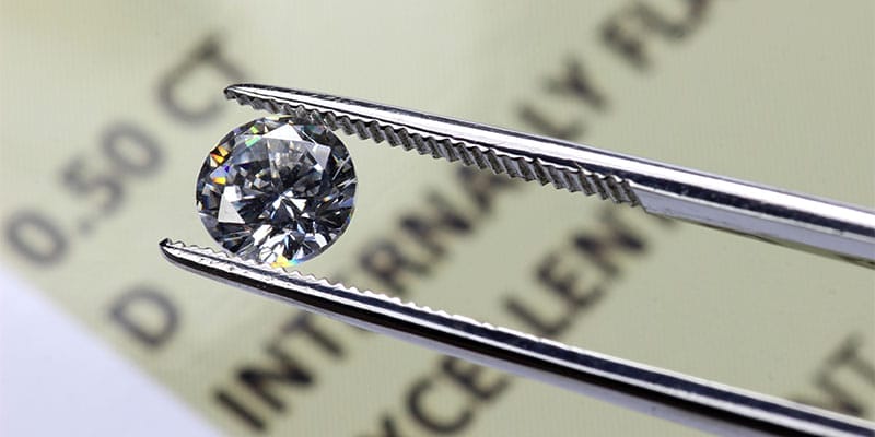 A cut diamond held by tweezers above an international diamond certificate.