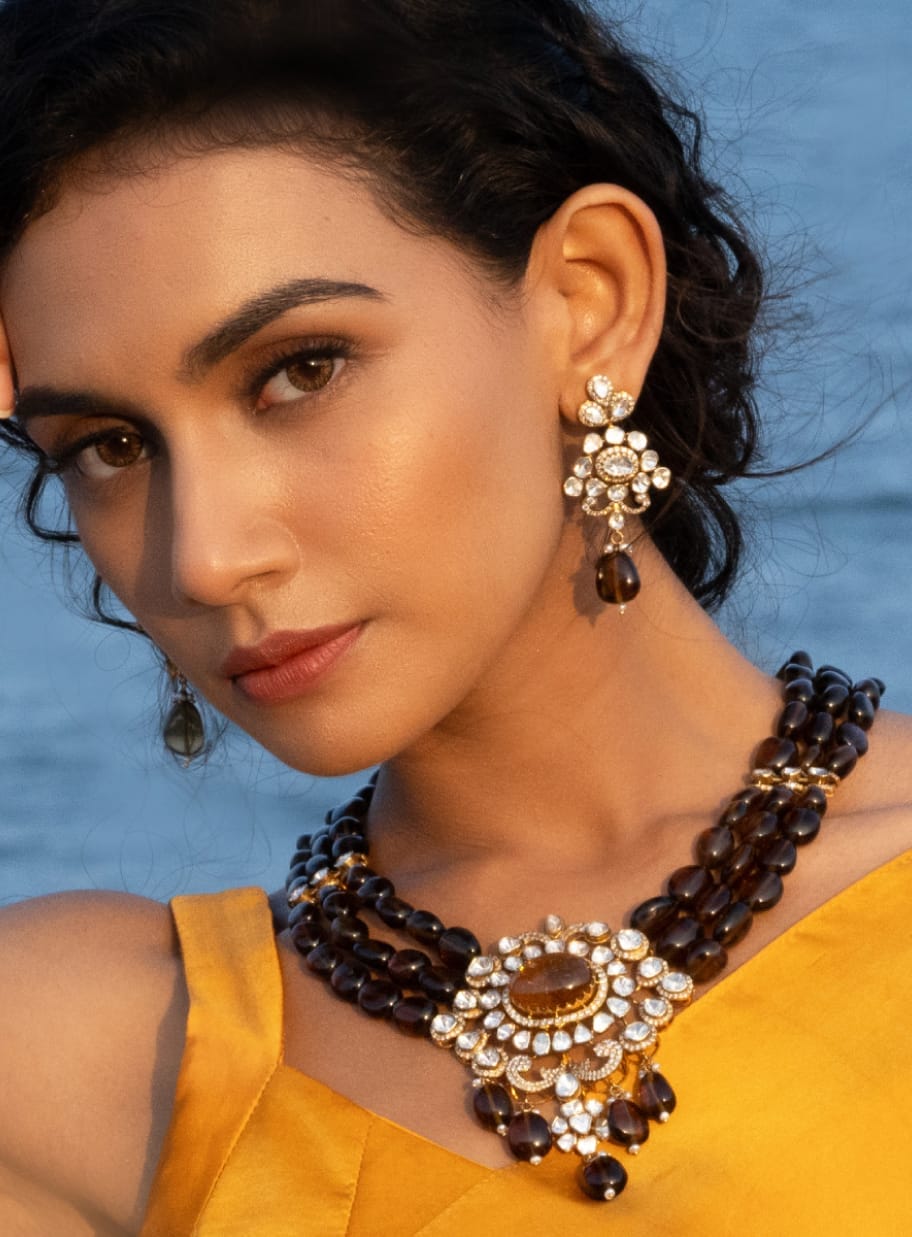 A close-up shot of a modern maid in a mustard color outfit adorning herself with stunning uncut diamond polki jewellery with precious gemstones from the Fazza Collection by Khwaahish.